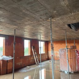 Grzegorz Kowalczyk GLOBAL-BUD - Wnętrze budynku w trakcie budowy z pustaków ceramicznych, z widocznym stropem podpartym stemplami. Na podłodze woda, w tle drabina i okna.