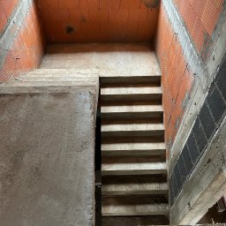 Grzegorz Kowalczyk GLOBAL-BUD - Surowe betonowe schody w surowym stanie, wewnątrz budynku z czerwonej cegły. Widok z góry na dół, podkreślający surowość i etap budowy.