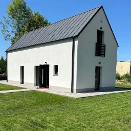 Nowoczesny dom jednorodzinny z szarą blachą na dachu, białą elewacją i otwartymi drzwiami wejściowymi. Widok z zewnątrz na zadbany trawnik i kostkę brukową.