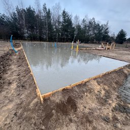 Grzegorz Kowalczyk GLOBAL-BUD - Zalany fundament pod budowę domu jednorodzinnego. Widoczne drewniane szalunki i wystające rury instalacyjne. W tle las i pochmurne niebo.