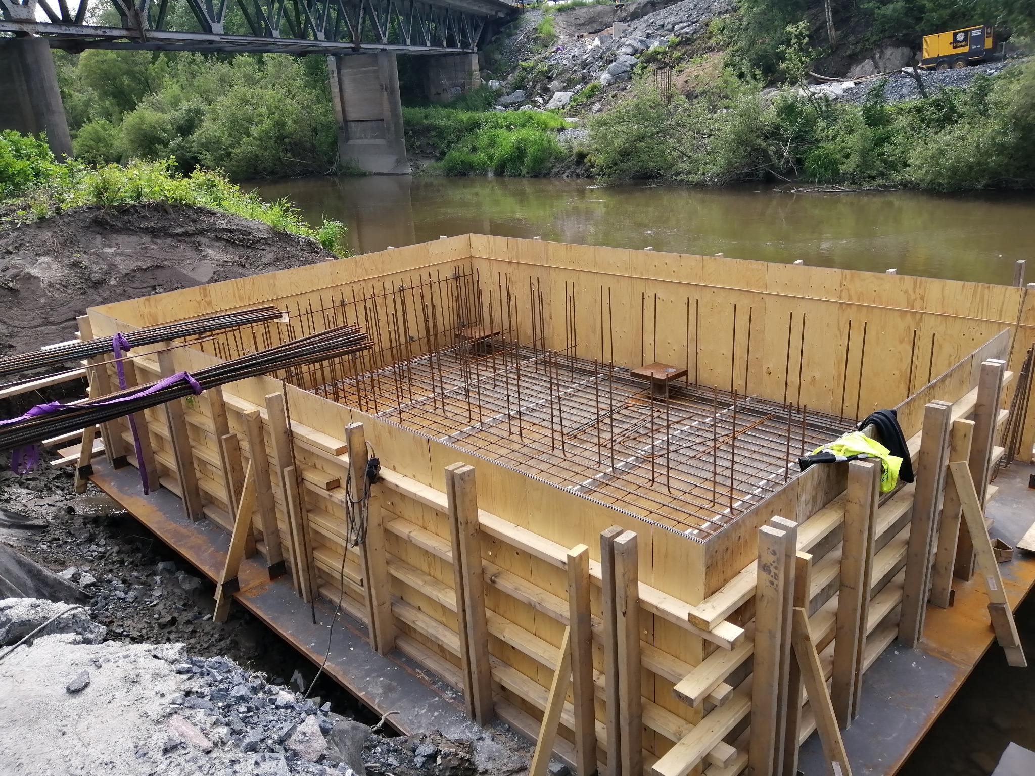 Szczegółowe ujęcie szalunku zbrojeniowego na fundamencie, z widoczną konstrukcją stalową i drewnianą formą, na tle rzeki i mostu. Prace ziemne w toku.