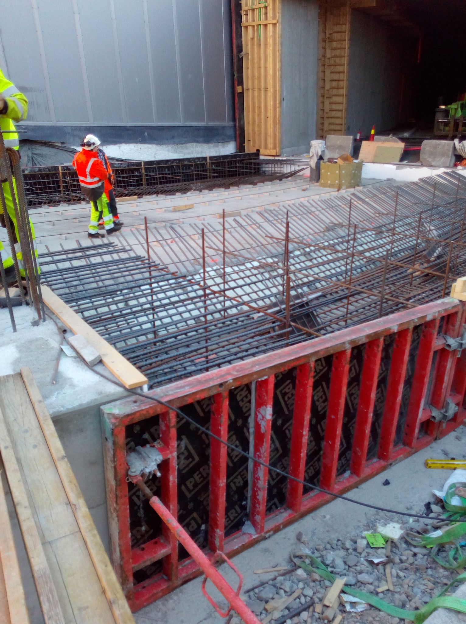 Zbrojenie fundamentów w czerwonym szalunku, widoczni pracownicy w odblaskowych ubraniach. Konstrukcja z metalowych prętów na budowie.