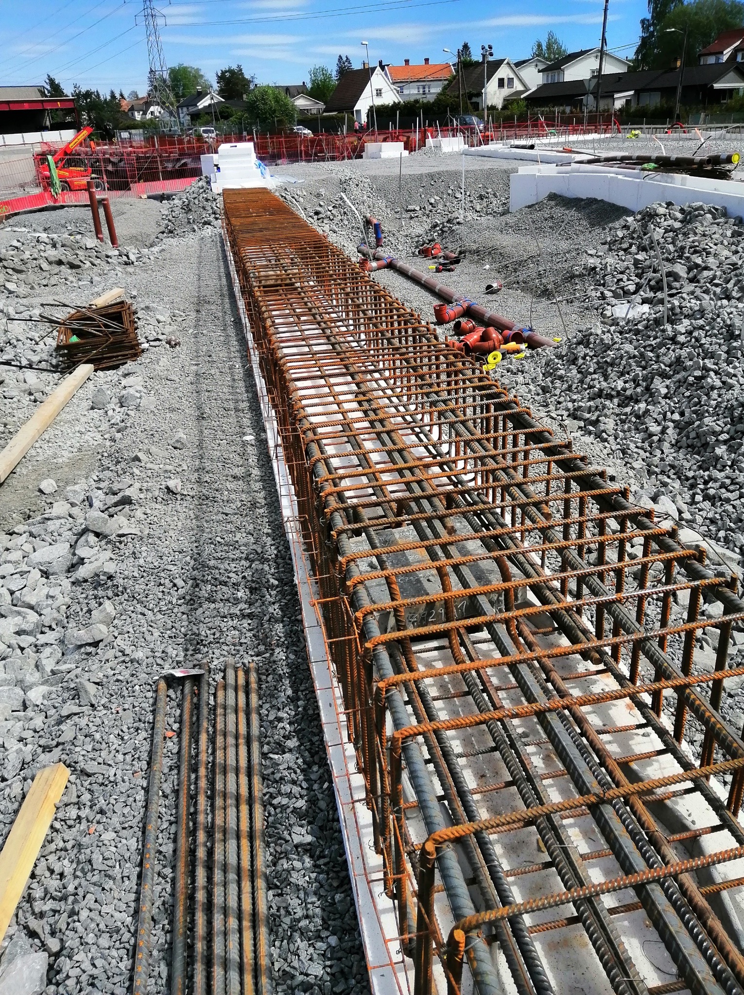 Zbrojenie fundamentów w wykopie, widok z góry. Stalowe pręty tworzą szkielet konstrukcji, przygotowany do zalania betonem. W tle gruz i elementy infrastruktury.