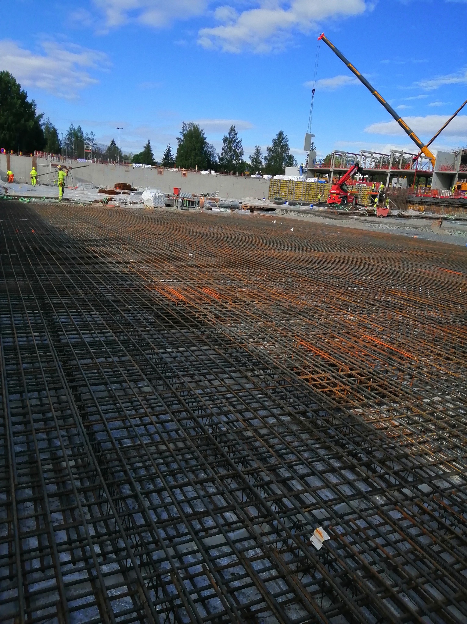 Szeroki kadr na rozległą siatkę zbrojeniową na budowie, z dźwigiem w tle i pracownikami w odblaskowych ubraniach. Niebo błękitne z chmurami.