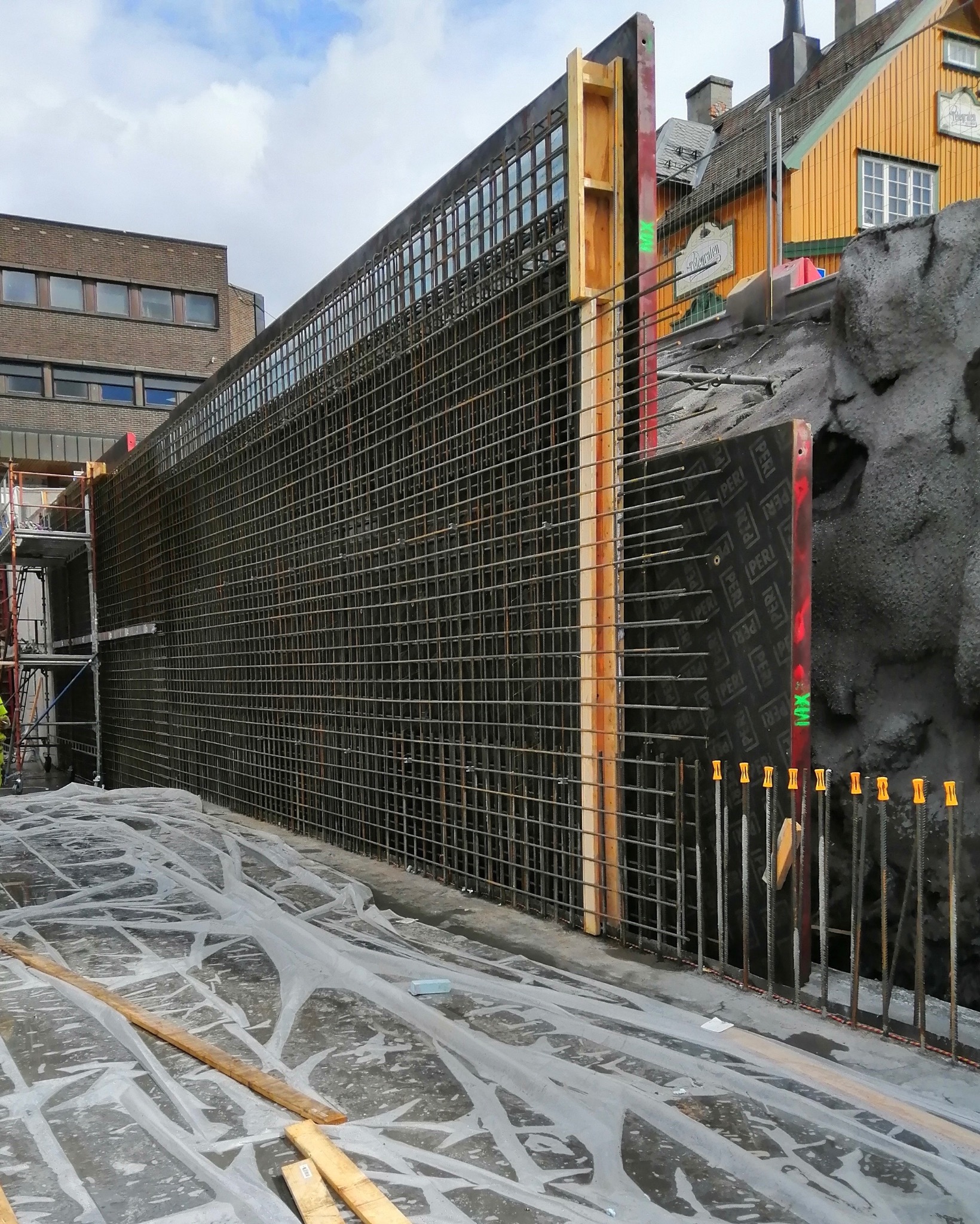 Siatka zbrojeniowa z drewnianym szalunkiem na tle miejskiej zabudowy. Widoczne elementy konstrukcyjne i zabezpieczenia na folii. Detal zbrojenia przed betonowaniem.