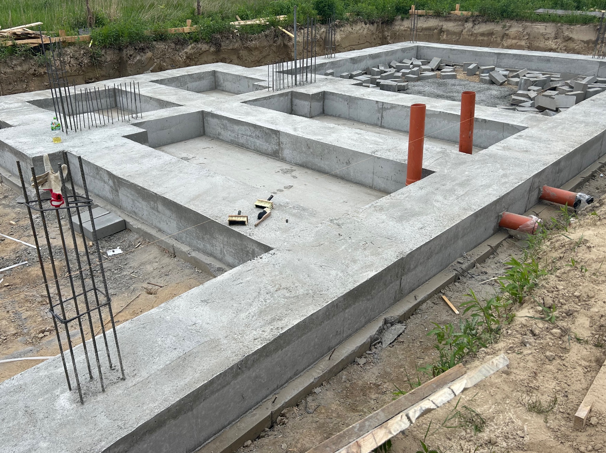 Zalane betonem fundamenty z wystającymi prętami zbrojeniowymi i pomarańczowymi rurami kanalizacyjnymi. Na fundamencie leżą bloczki betonowe.