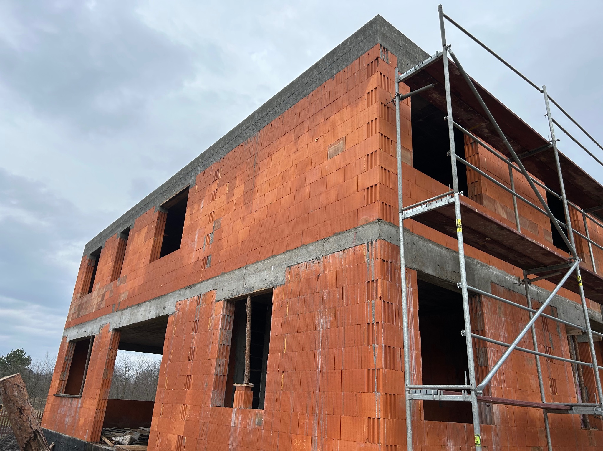 Surowy stan dwukondygnacyjnego domu z czerwonej cegły ceramicznej, z widocznymi otworami okiennymi i betonowymi nadprożami, otoczonego rusztowaniem.