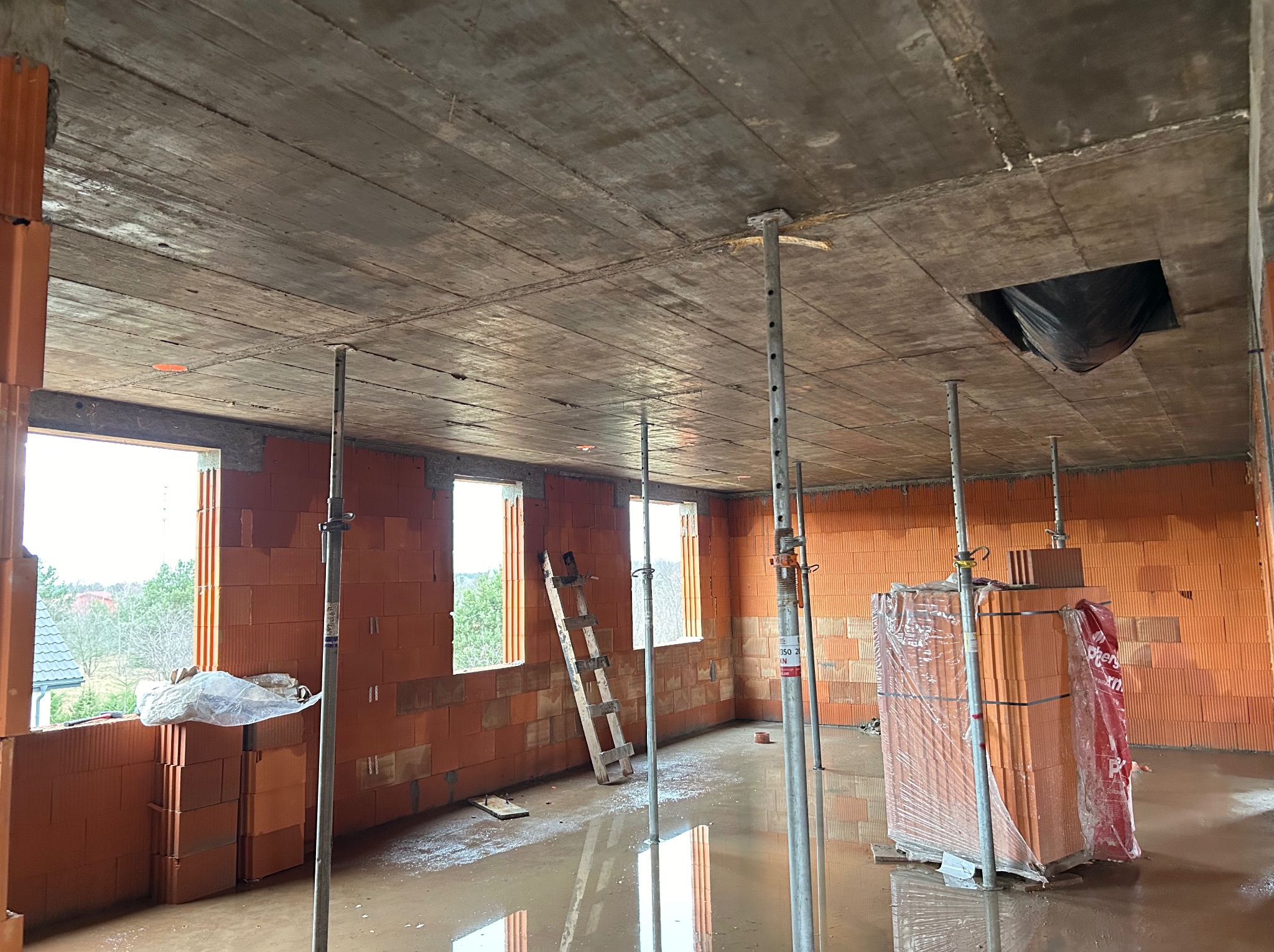 Wnętrze budynku w trakcie budowy z pustaków ceramicznych, z widocznym stropem podpartym stemplami. Na podłodze woda, w tle drabina i okna.