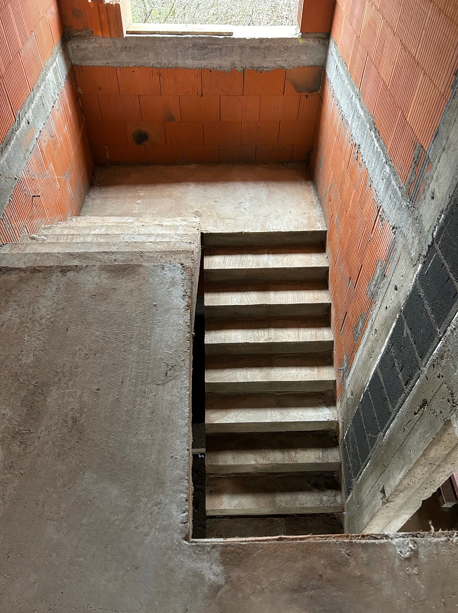 Surowe betonowe schody w surowym stanie, wewnątrz budynku z czerwonej cegły. Widok z góry na dół, podkreślający surowość i etap budowy.