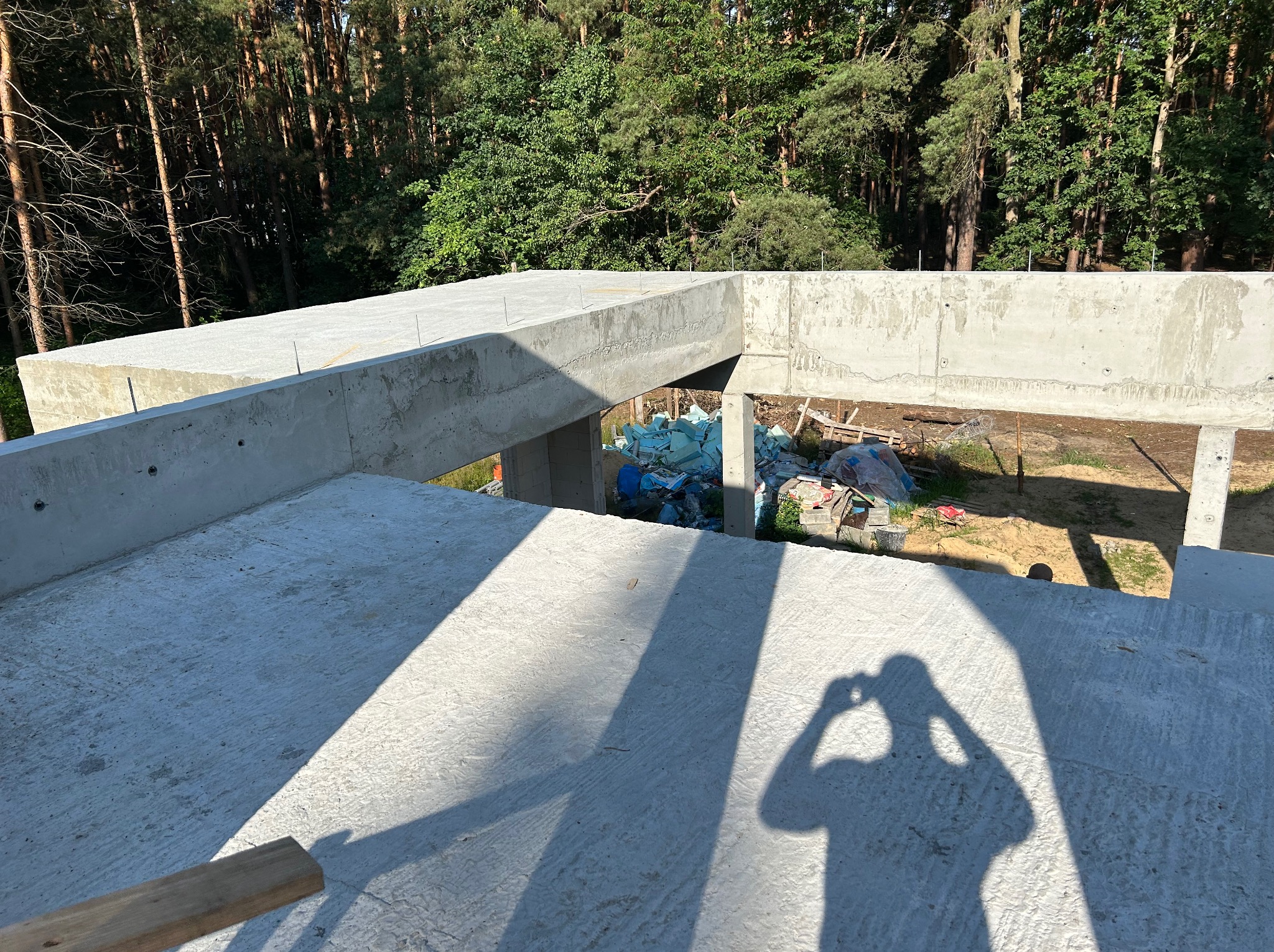 Surowa konstrukcja żelbetowa w lesie, cień fotografa na pierwszym planie. Widoczne szalunki i materiały budowlane. Ujęcie z góry, podkreślające detale betonu.