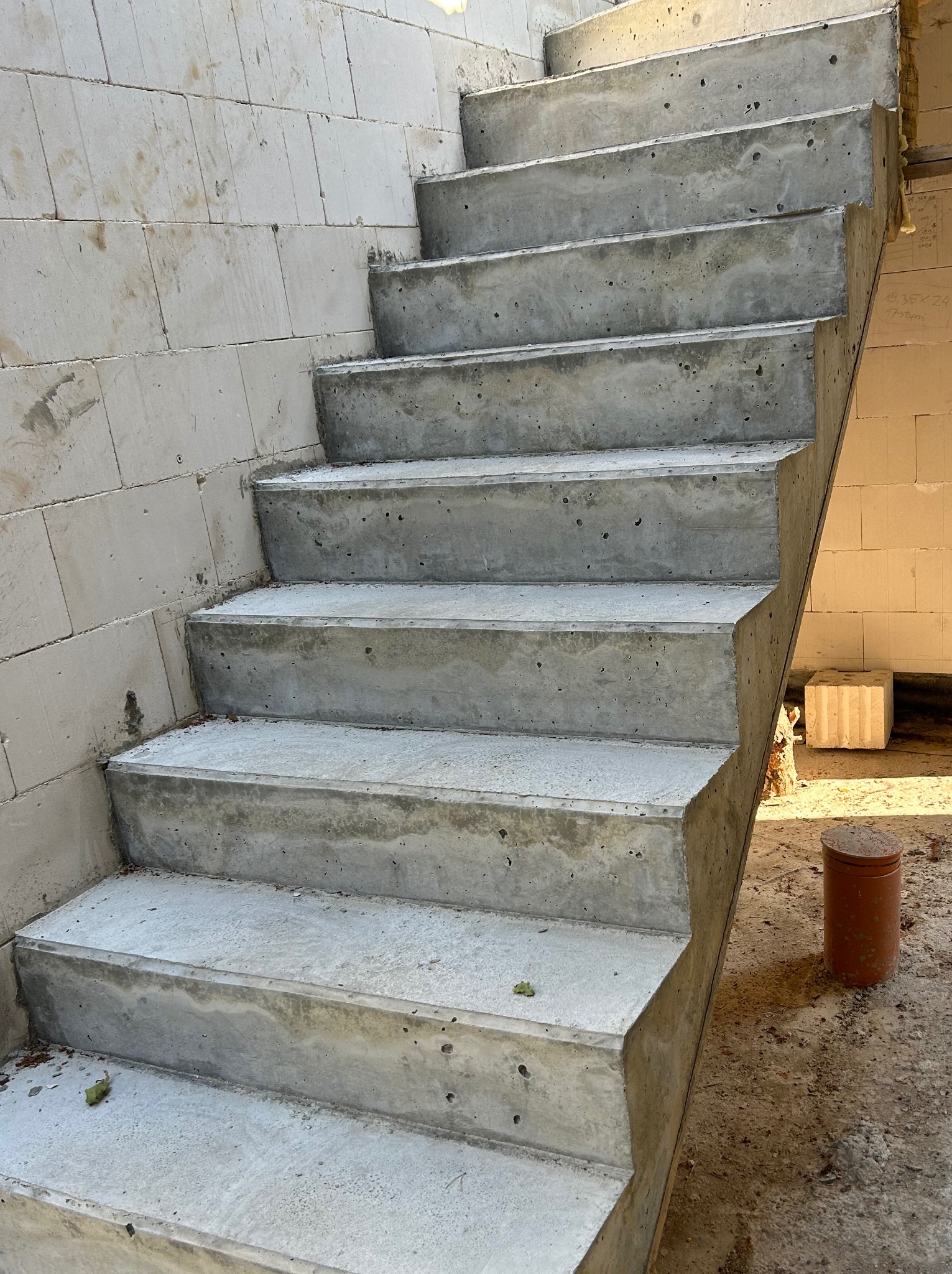 Betonowe schody w surowym stanie, widoczne ślady szalunku, ściany z bloczków betonowych, pomarańczowa rura na ziemi. Surowy stan budowy.