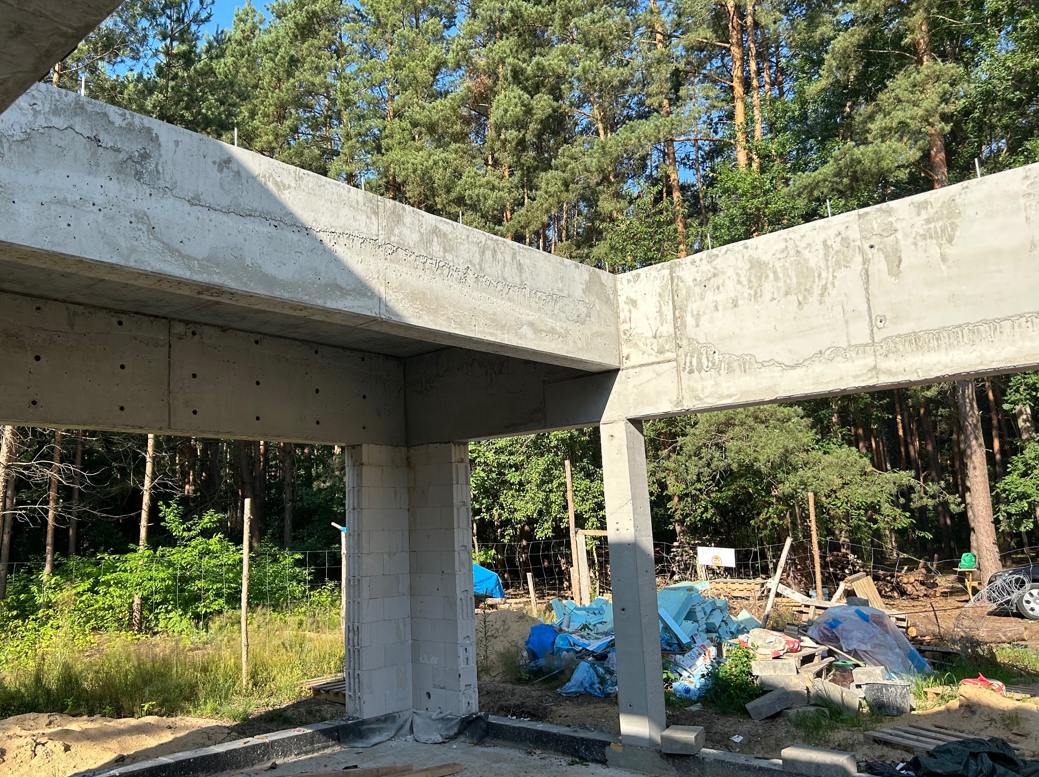 Surowa konstrukcja żelbetowa domu w lesie, widok z wnętrza. Szare ściany i filary na tle zieleni drzew. Elementy szalunków i materiałów budowlanych w tle.
