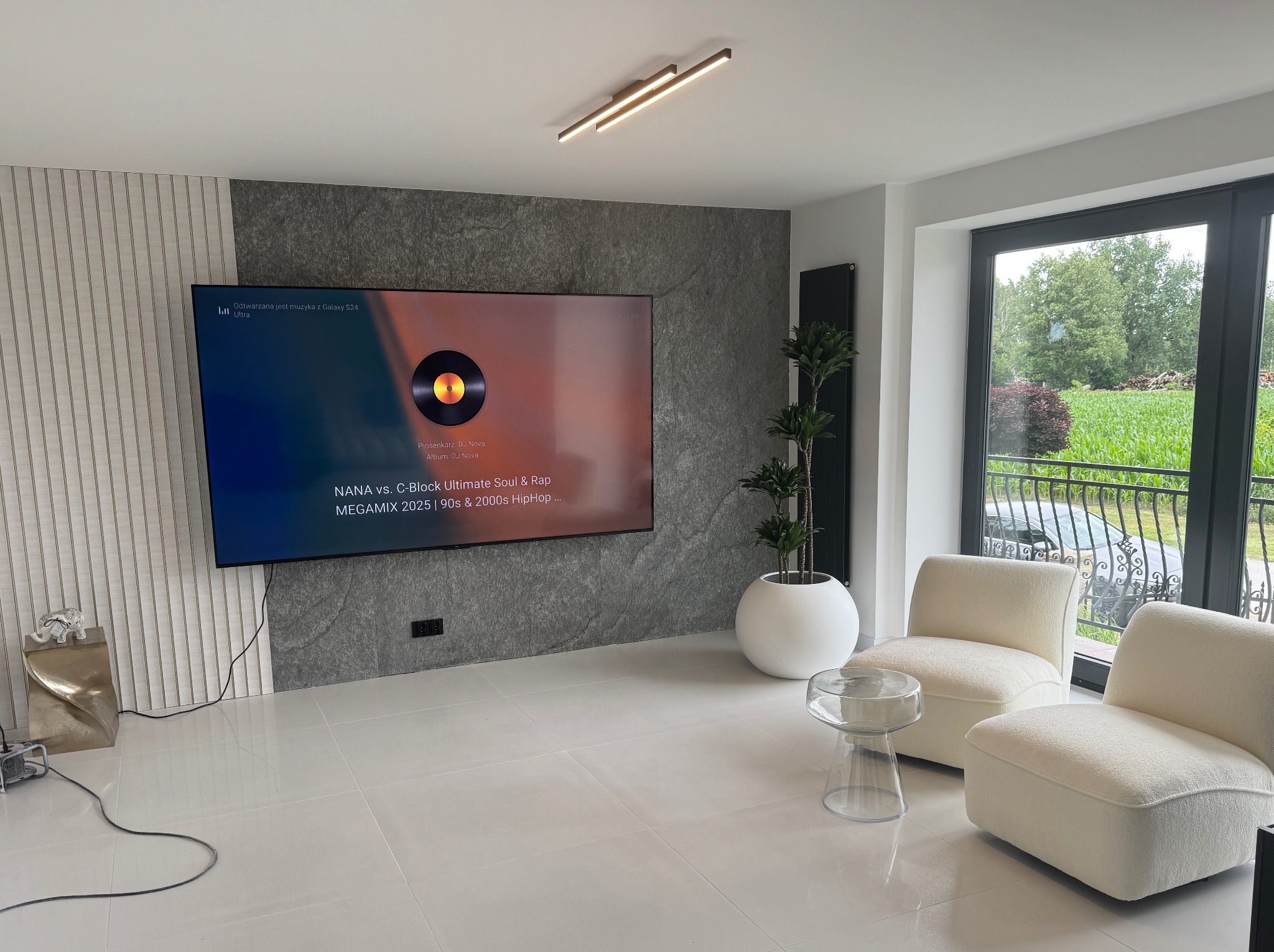 Nowoczesny salon z białymi płytkami podłogowymi, szarą ścianą z telewizorem, dwoma fotelami i rośliną w białej donicy. Widok na zieleń za oknem.