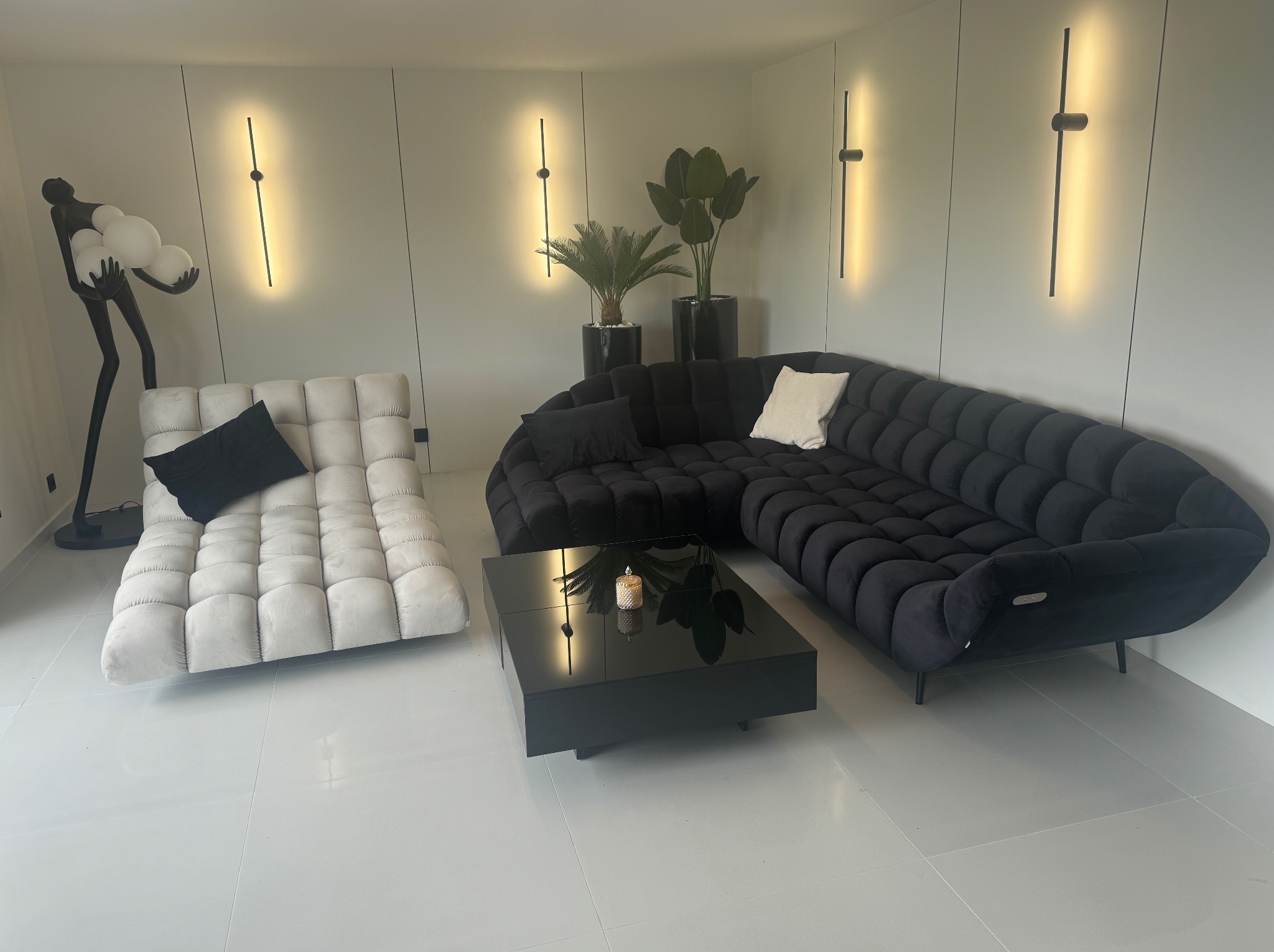 Nowoczesny salon z narożną sofą w kolorze antracytu, jasną leżanką i czarnym stolikiem kawowym. Minimalistyczne oświetlenie ścienne i rośliny doniczkowe dodają elegancji.