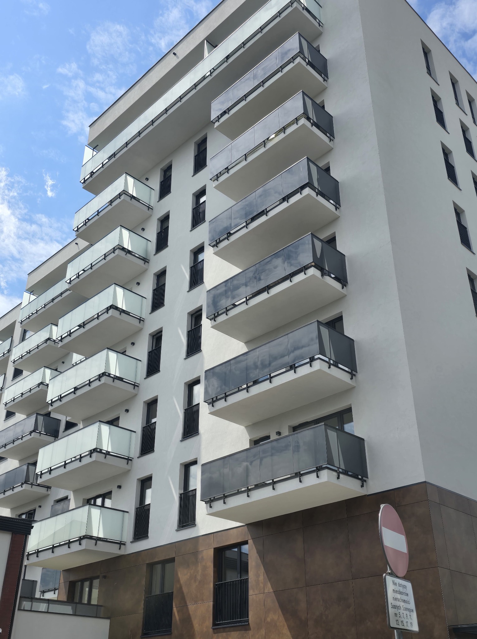 Nowoczesny budynek wielorodzinny z białymi ścianami i balkonami z szarymi balustradami. Ujęcie z dołu, widoczne błękitne niebo. Minimalistyczna architektura.