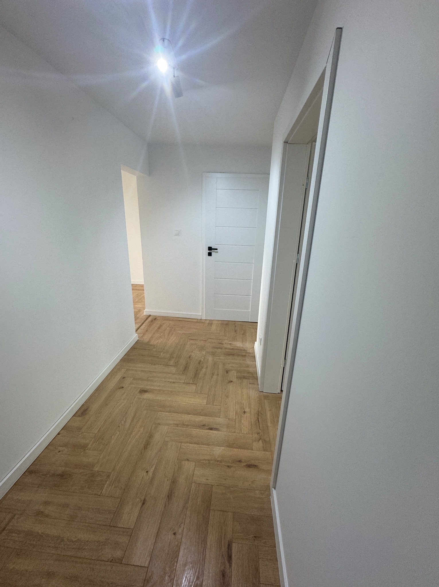 Jasne wnętrze korytarza z parkietem ułożonym w jodełkę. Białe ściany i drzwi tworzą minimalistyczny, nowoczesny wygląd. Widoczne oświetlenie sufitowe.