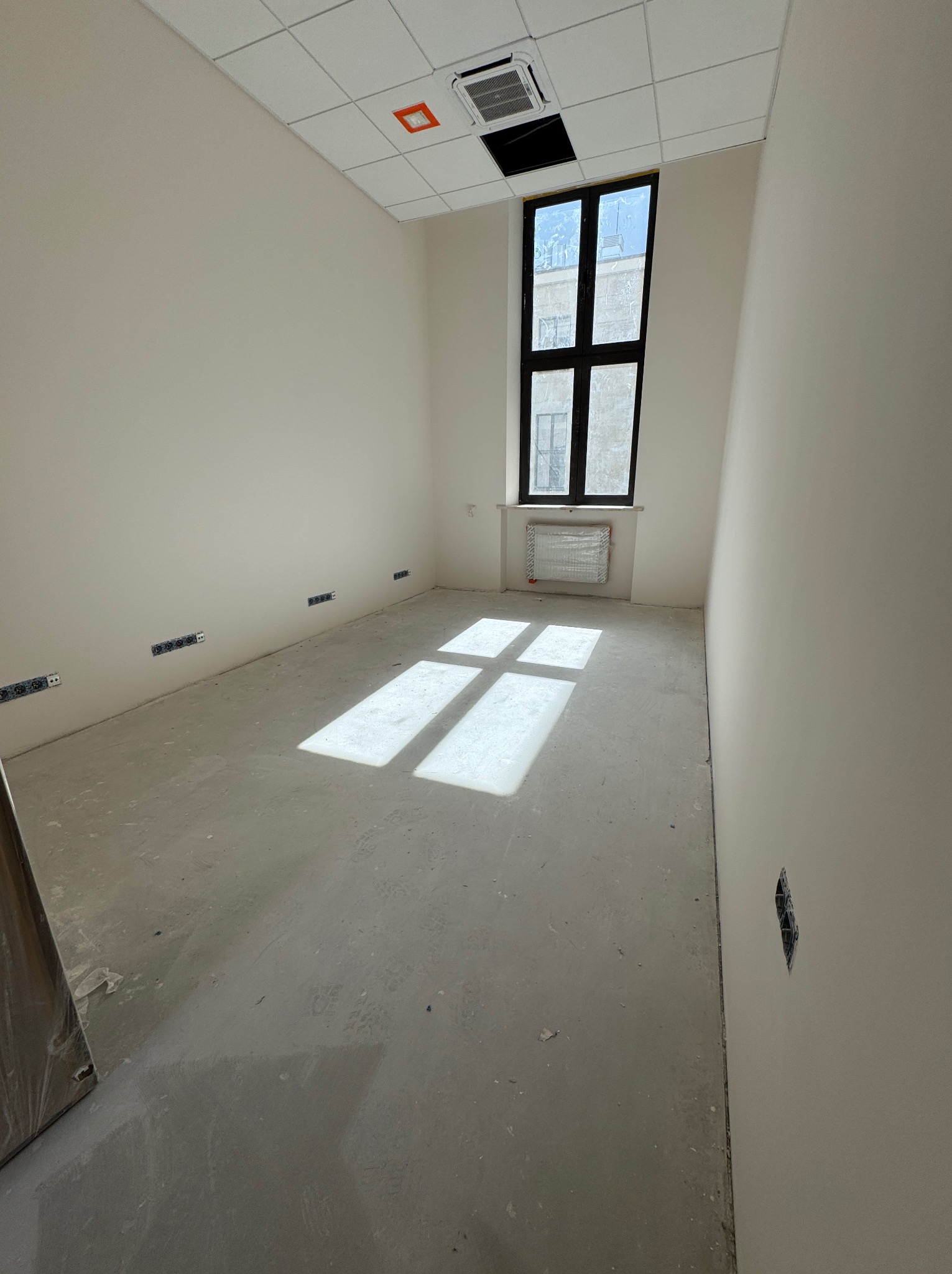 Wnętrze pokoju w stanie deweloperskim z oknem, grzejnikiem i widocznymi gniazdkami elektrycznymi. Na podłodze widoczne ślady po pracach budowlanych.