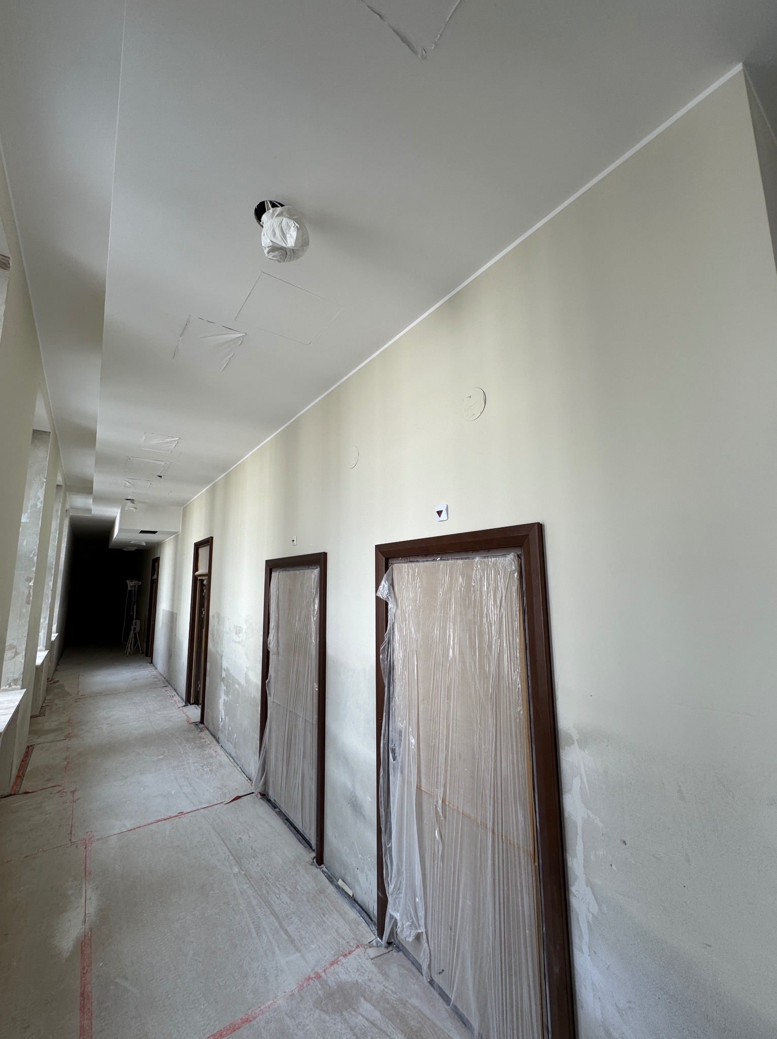 Korytarz w trakcie remontu: zabezpieczone folią drzwi, surowy beton na podłodze, ściany przygotowane do malowania. Widoczne ślady po pracach budowlanych.