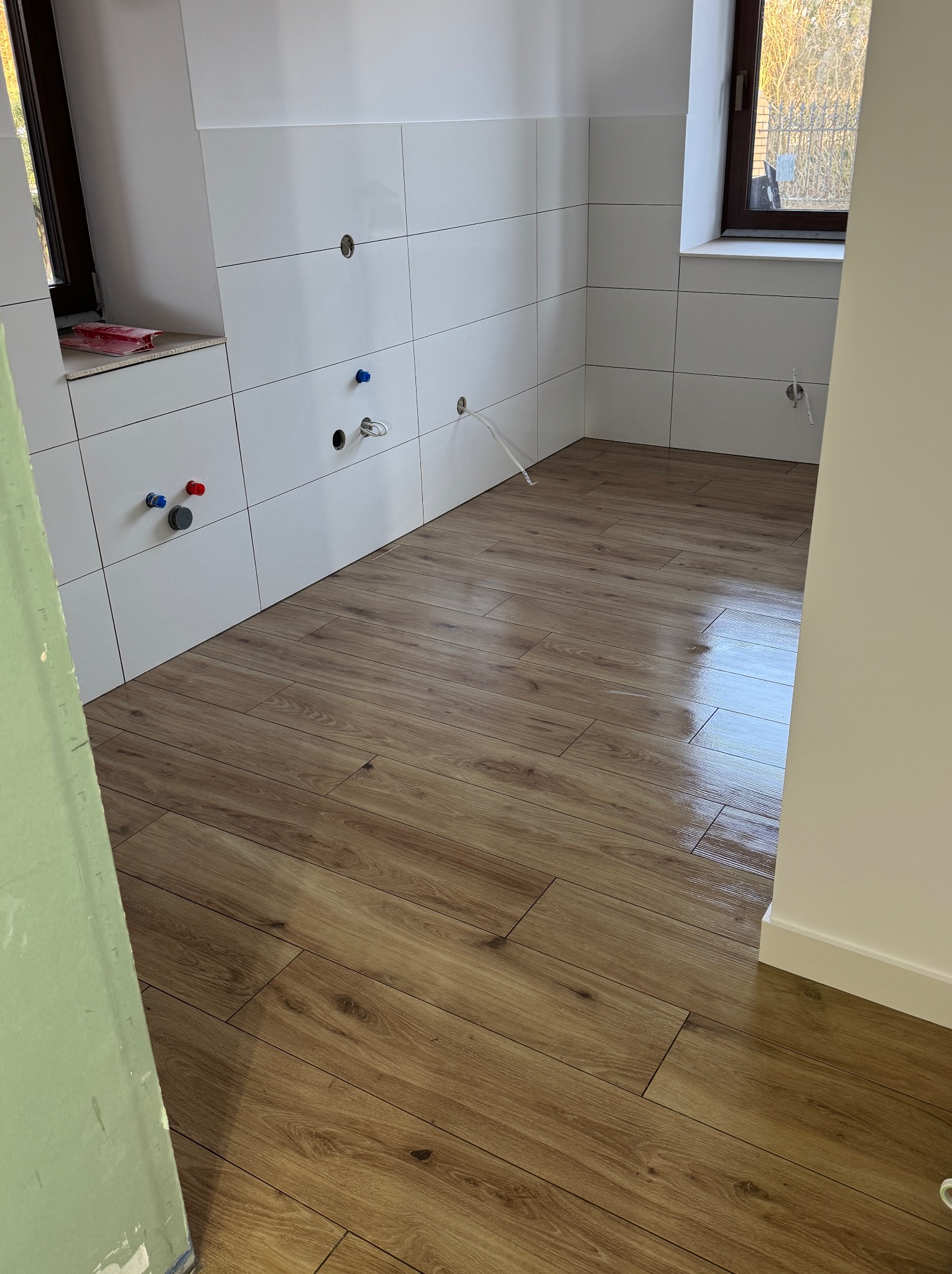 Wykończona łazienka z drewnopodobną podłogą i białymi płytkami ściennymi. Widoczne instalacje wodne i okno. Fragment ściany w kolorze złamanej bieli.