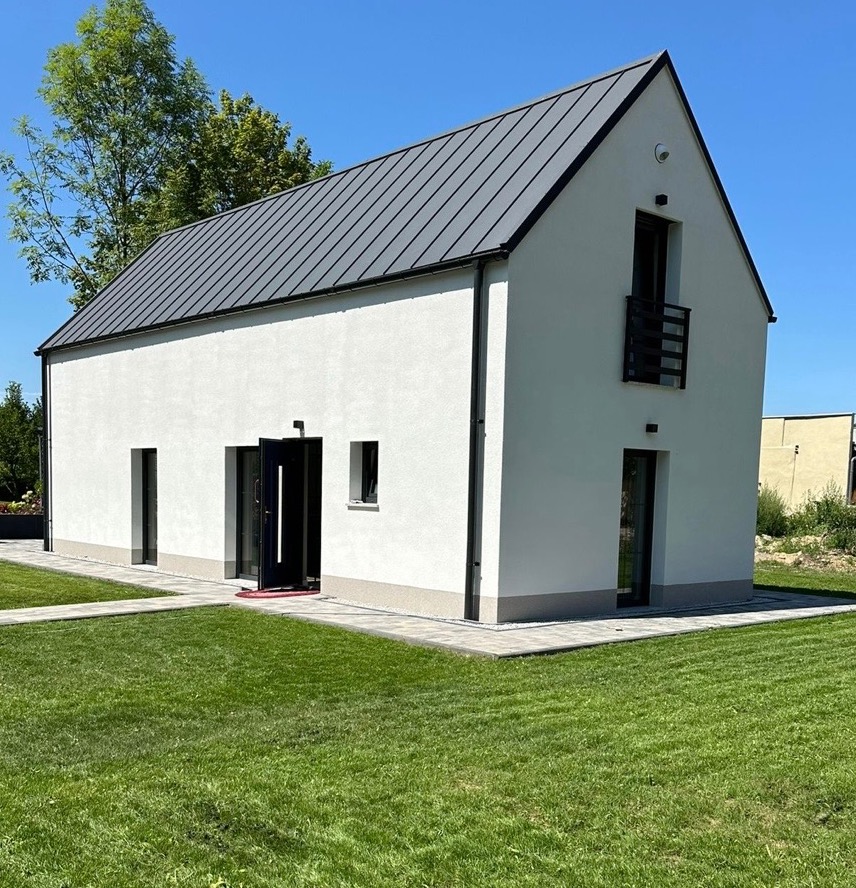 Nowoczesny dom jednorodzinny z szarą blachą na dachu, białą elewacją i otwartymi drzwiami wejściowymi. Widok z zewnątrz na zadbany trawnik i kostkę brukową.