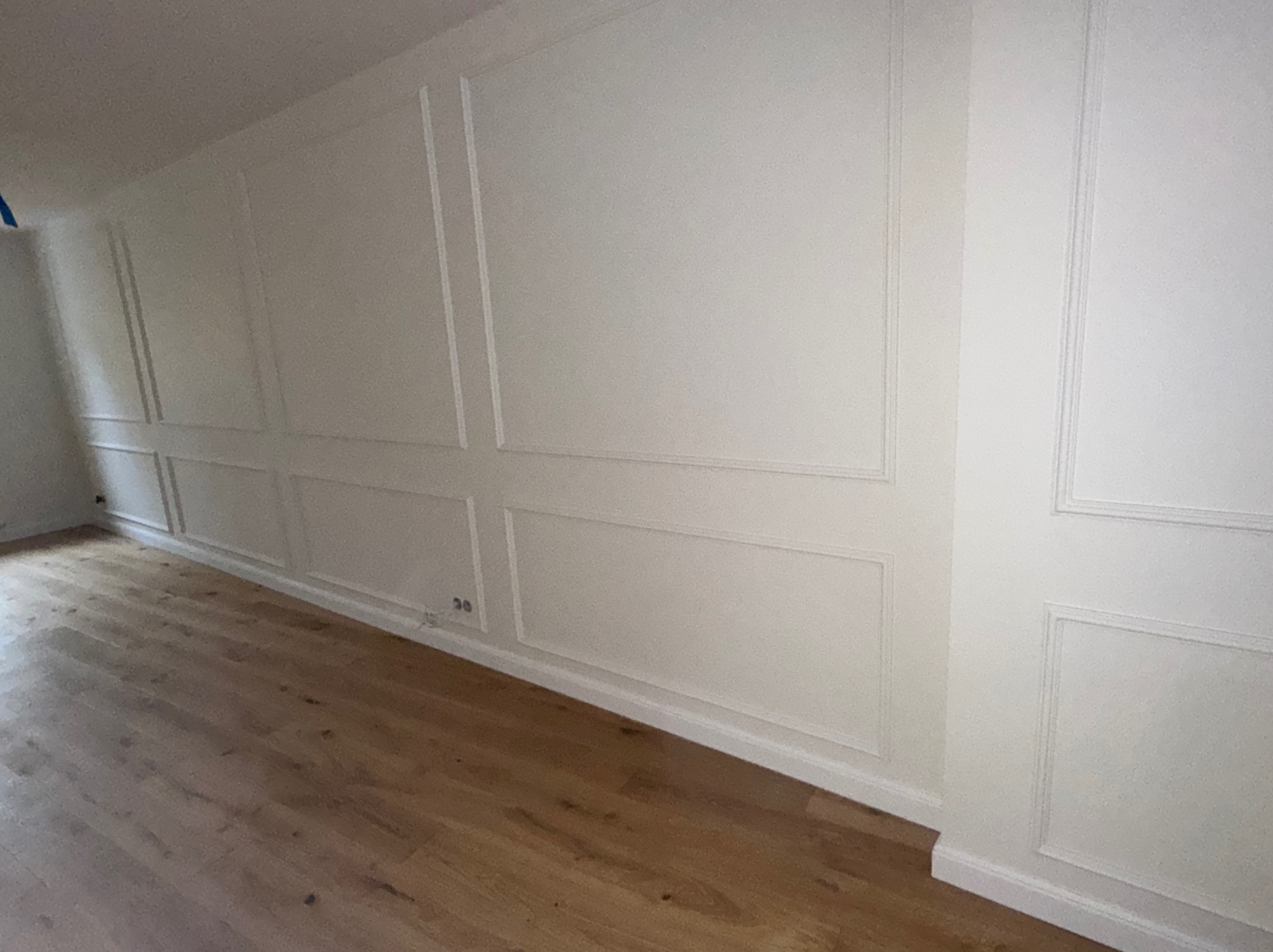 Biała ściana z dekoracyjnymi panelami i drewnianą podłogą. Widoczne gniazdka elektryczne przy podłodze. Ujęcie z perspektywy podłogi, podkreślające detale wykończenia.