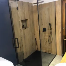 Nowoczesna kabina prysznicowa ze szklanymi ścianami i czarnymi profilami, drewnopodobne płytki na ścianach, czarna armatura i brodzik.