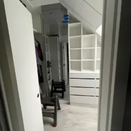 Wnętrze garderoby w skosie z białymi regałami i szufladami, widoczne przez otwarte drzwi, z drabinką w środku.