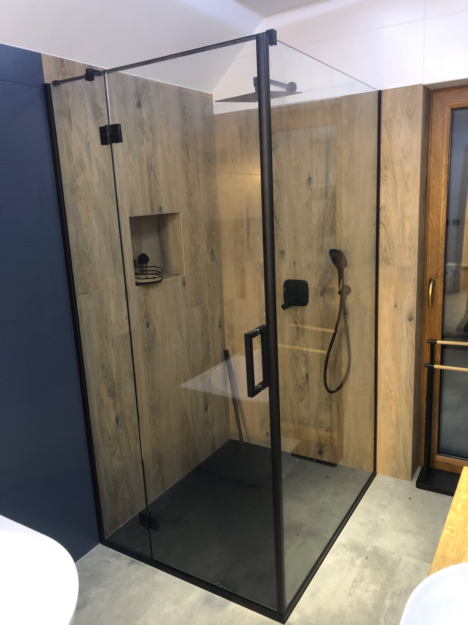 Nowoczesna kabina prysznicowa ze szkła z czarnymi profilami, drewnopodobne płytki na ścianach, widoczna słuchawka prysznicowa i półka na kosmetyki.