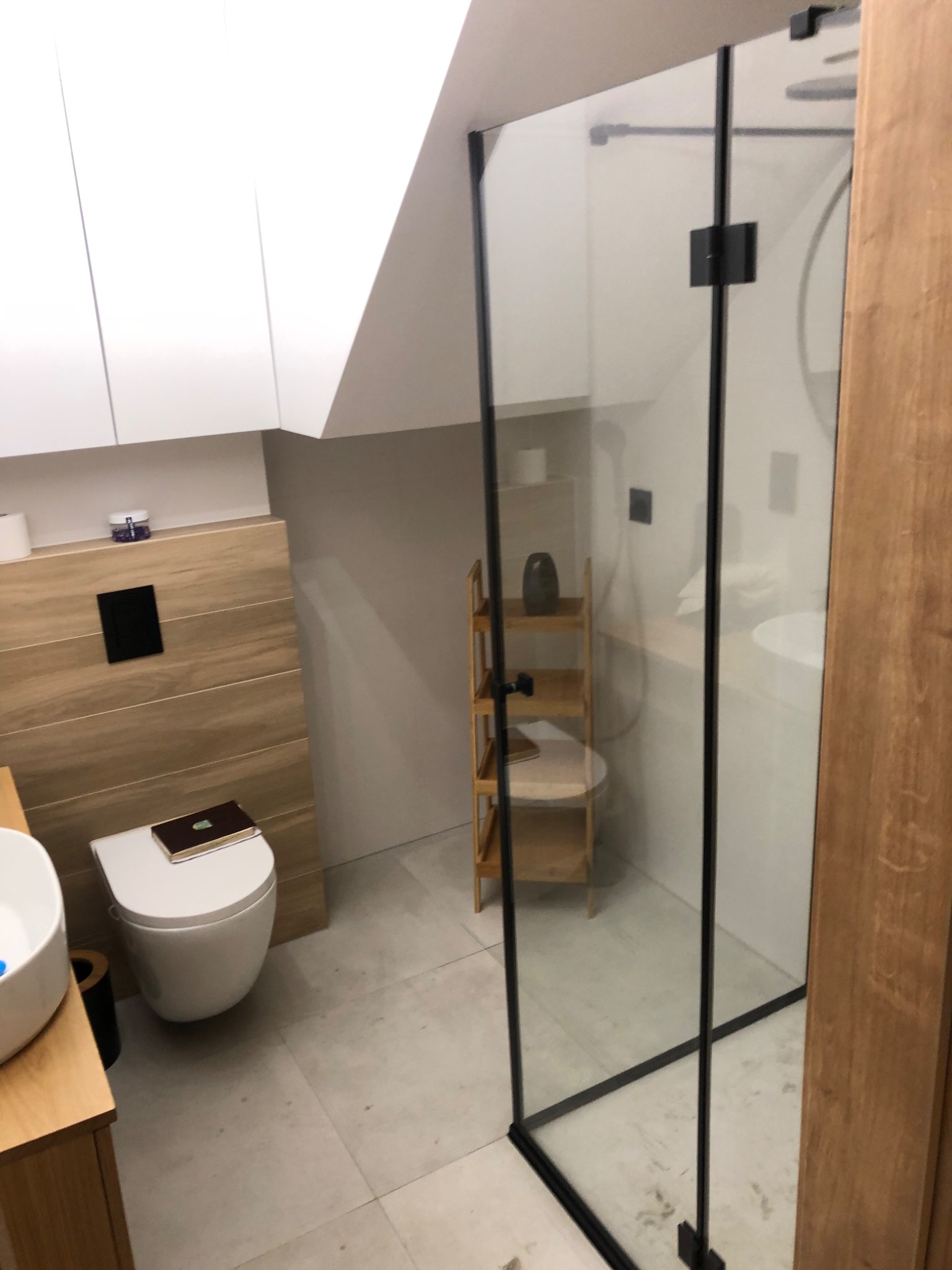 Nowoczesna łazienka na poddaszu z kabiną prysznicową ze szklanymi ściankami z czarnymi ramami, drewnianą zabudową toalety i umywalki, oraz półką z bambusa.