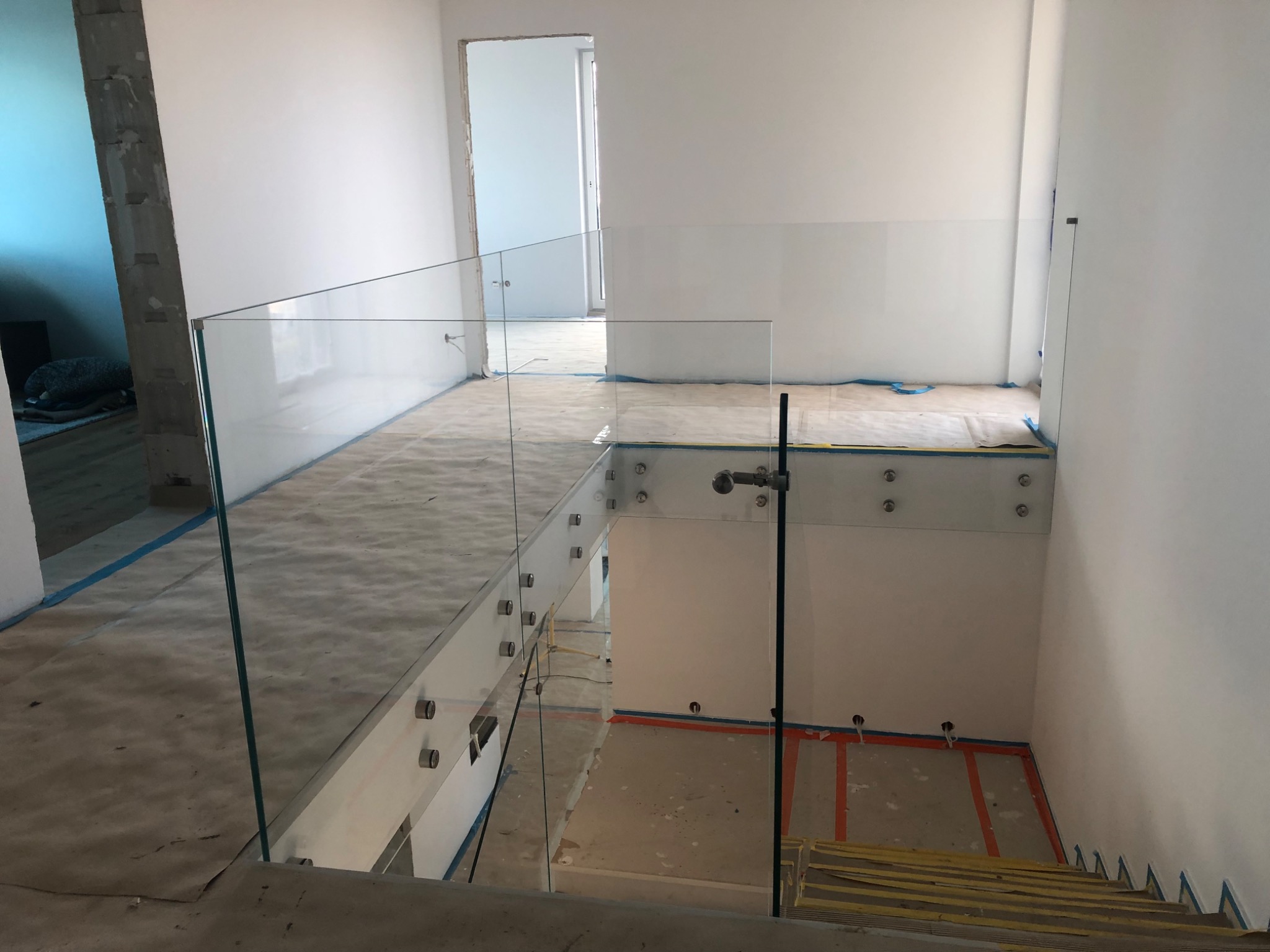 Wnętrze w trakcie wykańczania z zamontowaną szklaną balustradą na schodach i antresoli, widoczne metalowe mocowania i zabezpieczenia podłogi.