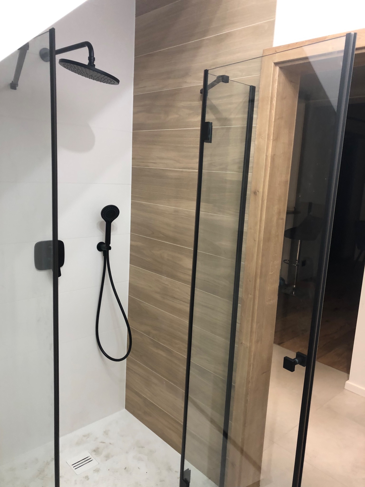 Nowoczesna kabina prysznicowa ze szklaną ścianką i czarną armaturą, widok z zewnątrz ukazujący fragment łazienki z drewnianą ramą drzwi i krzesłem barowym w tle.