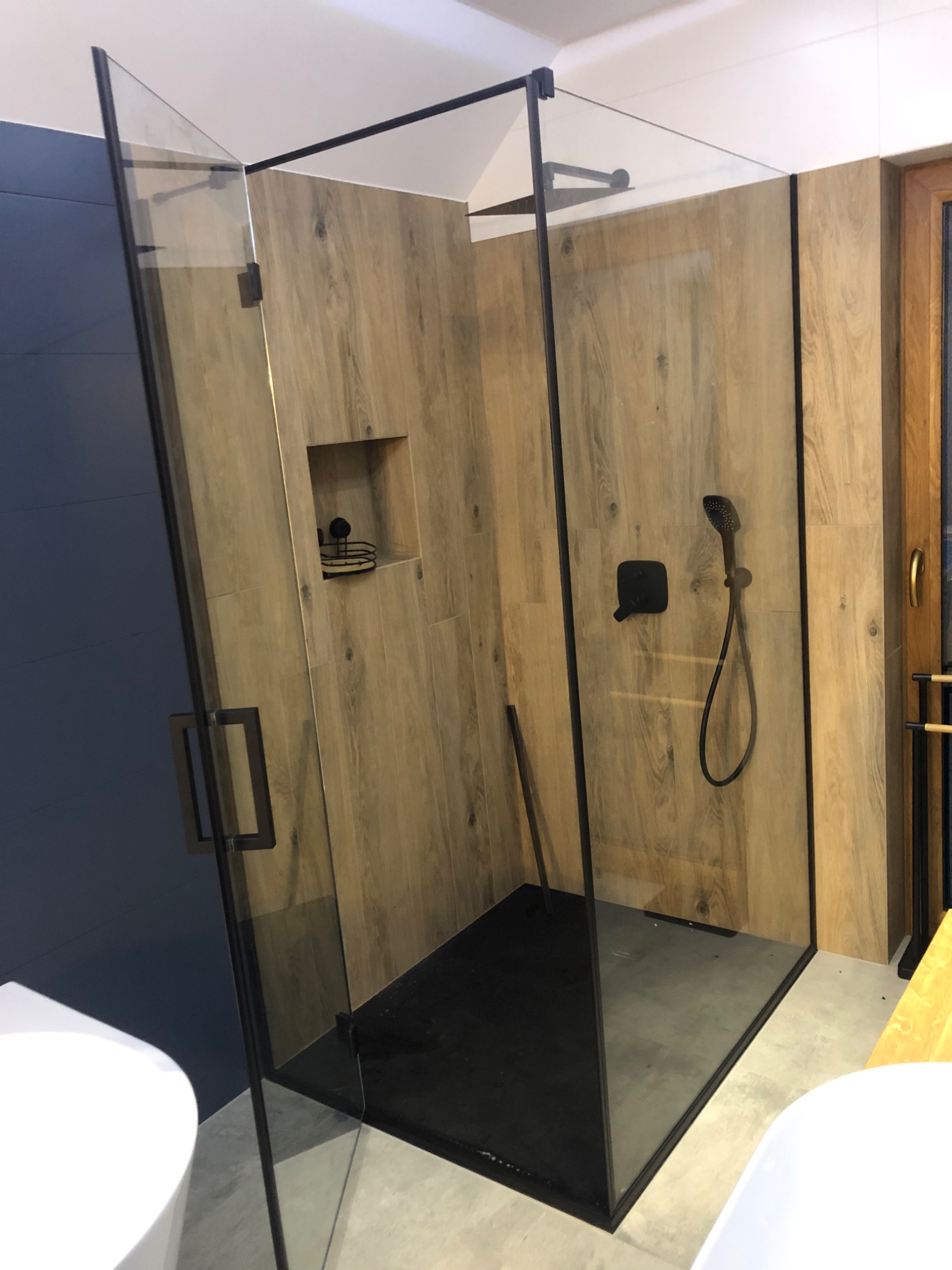 Nowoczesna kabina prysznicowa ze szklanymi ścianami i czarnymi profilami, drewnopodobne płytki na ścianach, czarna armatura i brodzik.