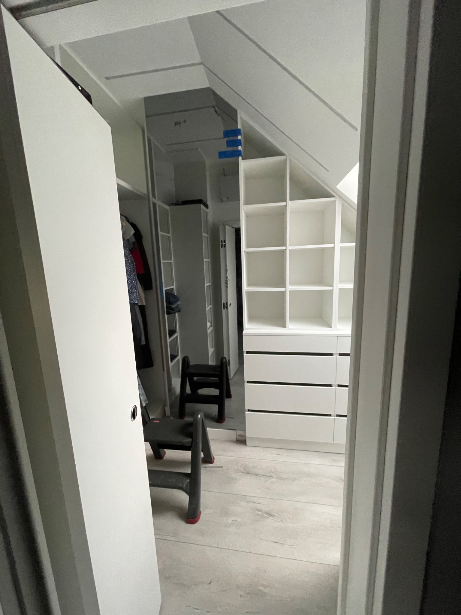 Wnętrze garderoby w skosie z białymi regałami i szufladami, widoczne przez otwarte drzwi, z drabinką w środku.