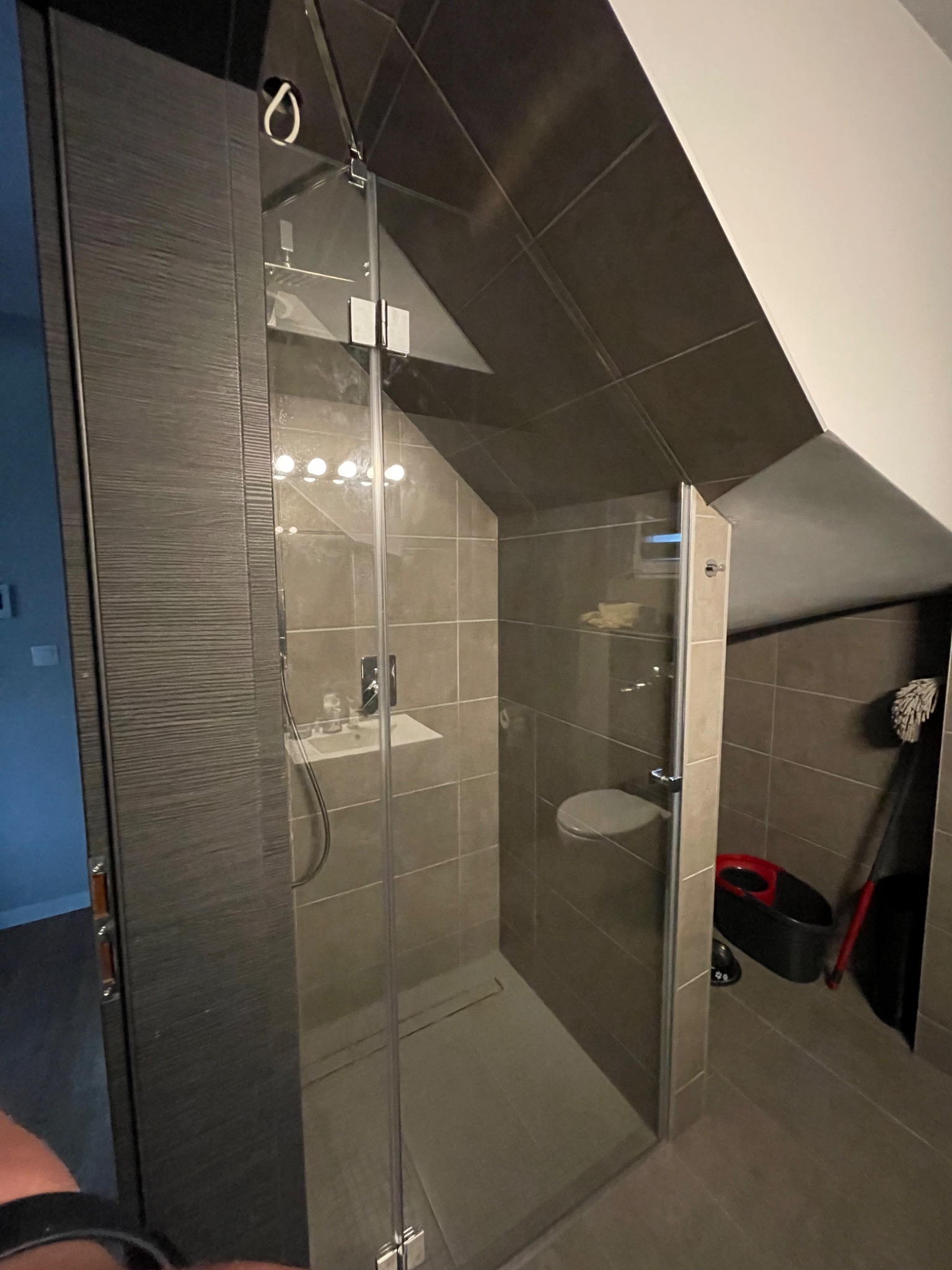 Szklana kabina prysznicowa w łazience na poddaszu z szarymi płytkami, widoczny mop i wiadro.