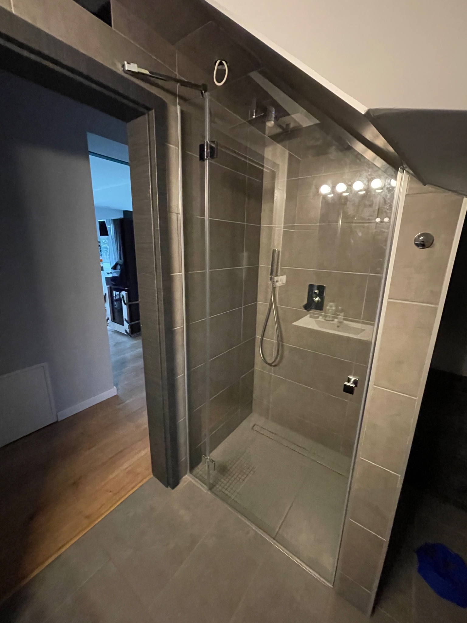 Nowoczesna kabina prysznicowa z szarą glazurą, widoczna przez otwarte drzwi do łazienki, z widocznym odpływem liniowym i minimalistyczną armaturą.