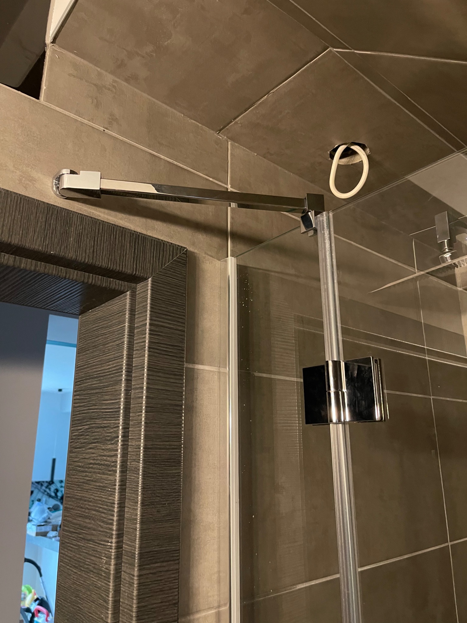 Szklana kabina prysznicowa z chromowanymi okuciami, widoczny uchwyt stabilizujący i fragment ściany z ciemnymi płytkami oraz element ramy drzwi z dekoracyjnym frezowaniem.