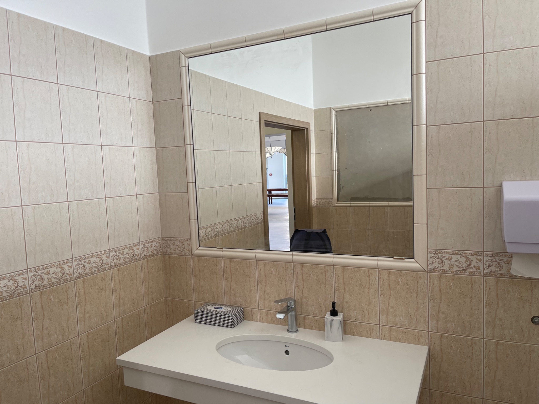 Prostokątne lustro w ramie nad umywalką w łazience wyłożonej beżowymi płytkami z dekoracyjnym pasem, odbijające fragment pomieszczenia z widocznym przejściem do innego pokoju.