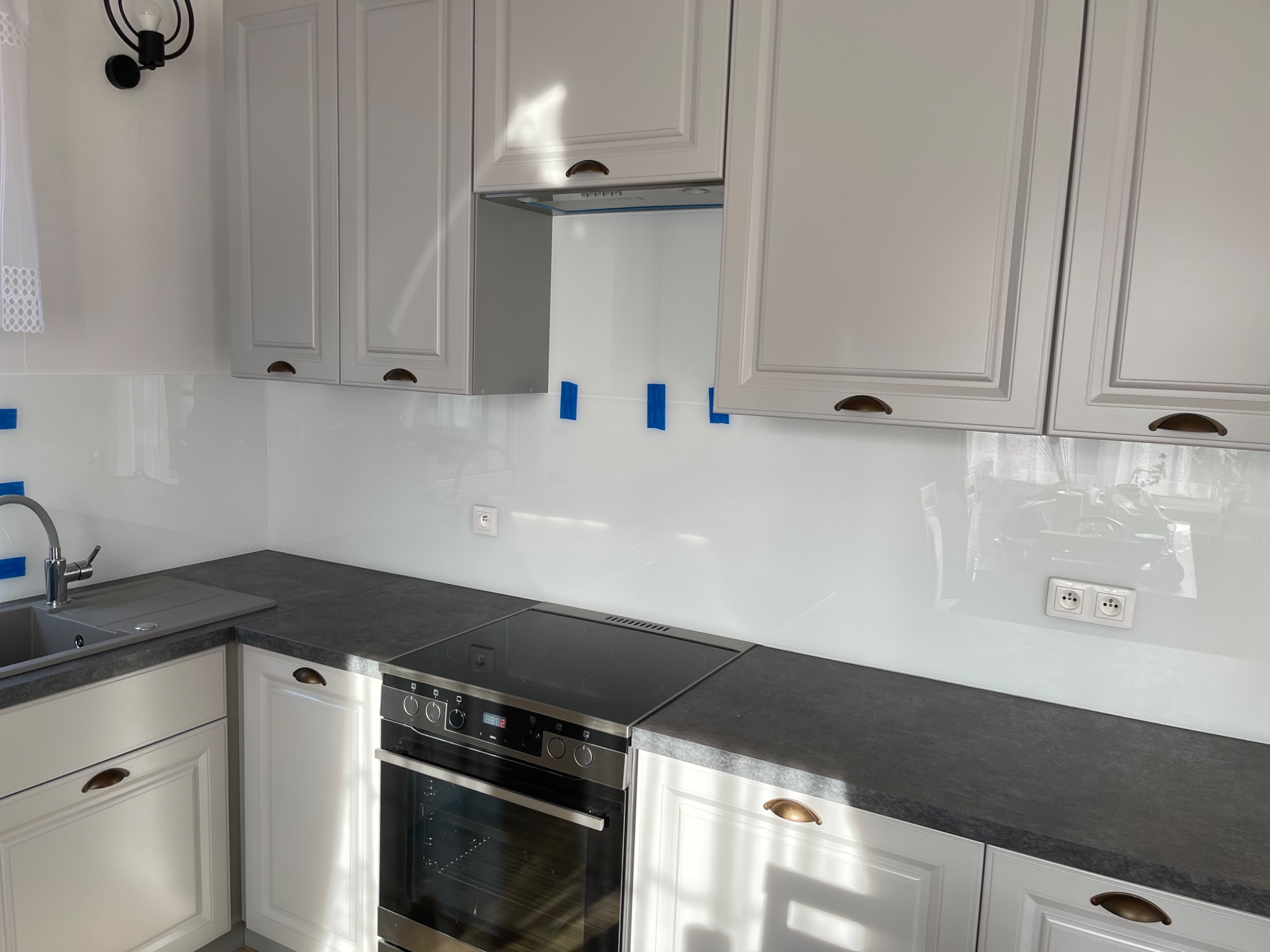 Biała szklana ściana nad blatem kuchennym z szarym blatem, widoczne gniazdka elektryczne i niebieska taśma montażowa, sugerująca świeży montaż.