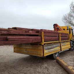 Żółta ciężarówka z dźwigiem załadowana świeżo przetartym drewnem w odcieniach czerwieni, widok z boku na placu składowym z pniem drzewa w pierwszym planie i budynkiem w tle.