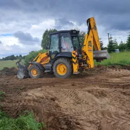 Żółto-czarna koparko-ładowarka JCB na nierównym, błotnistym terenie z widocznymi śladami opon, w tle zielona łąka i pochmurne niebo.