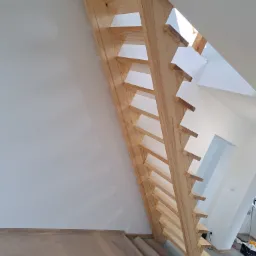 Nowoczesne, drewniane schody samonośne z jasnego drewna, widok od dołu, minimalistyczny design, białe ściany, jasne wnętrze, częściowo zakryte podłogi zabezpieczone kartonem.