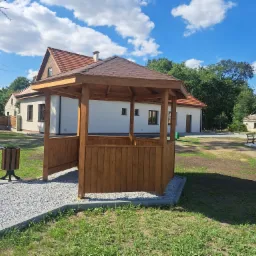 Nowo wybudowana, drewniana altana z brązowym dachem, otoczona zielonym trawnikiem i ścieżką z jasnego żwiru, na tle budynków z czerwonymi dachami pod błękitnym niebem z białymi obłokami.