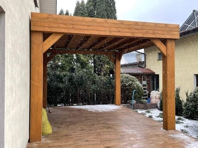 Drewniana konstrukcja z belek w Gostyninie, prawdopodobnie zadaszenie tarasu lub wiata, pokryta delikatnym śniegiem; widoczne drewniane słupy i belkowanie, otoczenie z elementami ogrodu i zabudowy...