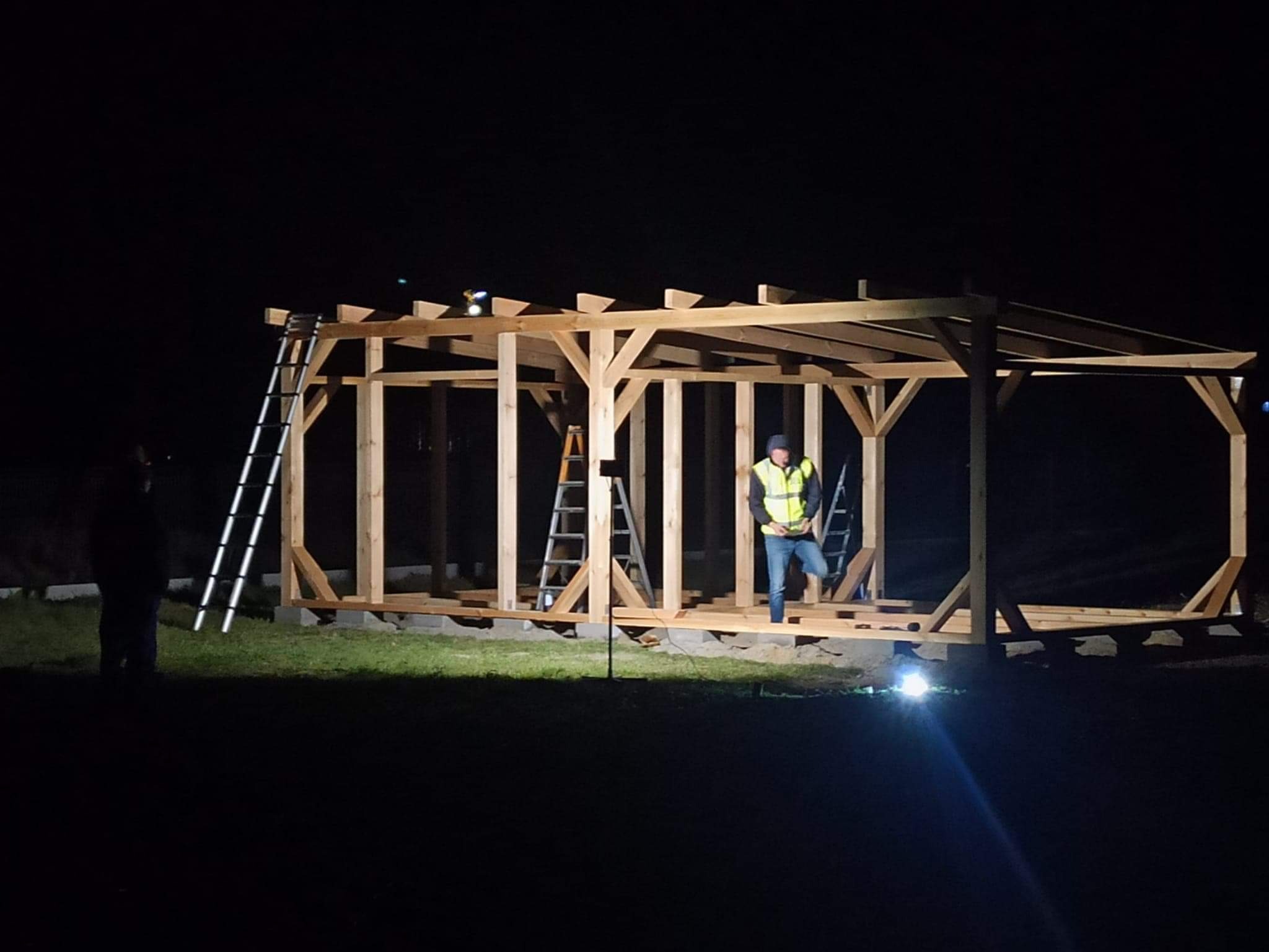 Nocna fotografia w trakcie budowy drewnianej konstrukcji altany, widoczny pracownik w kamizelce odblaskowej i drabiny, oświetlenie halogenem budowlanym.