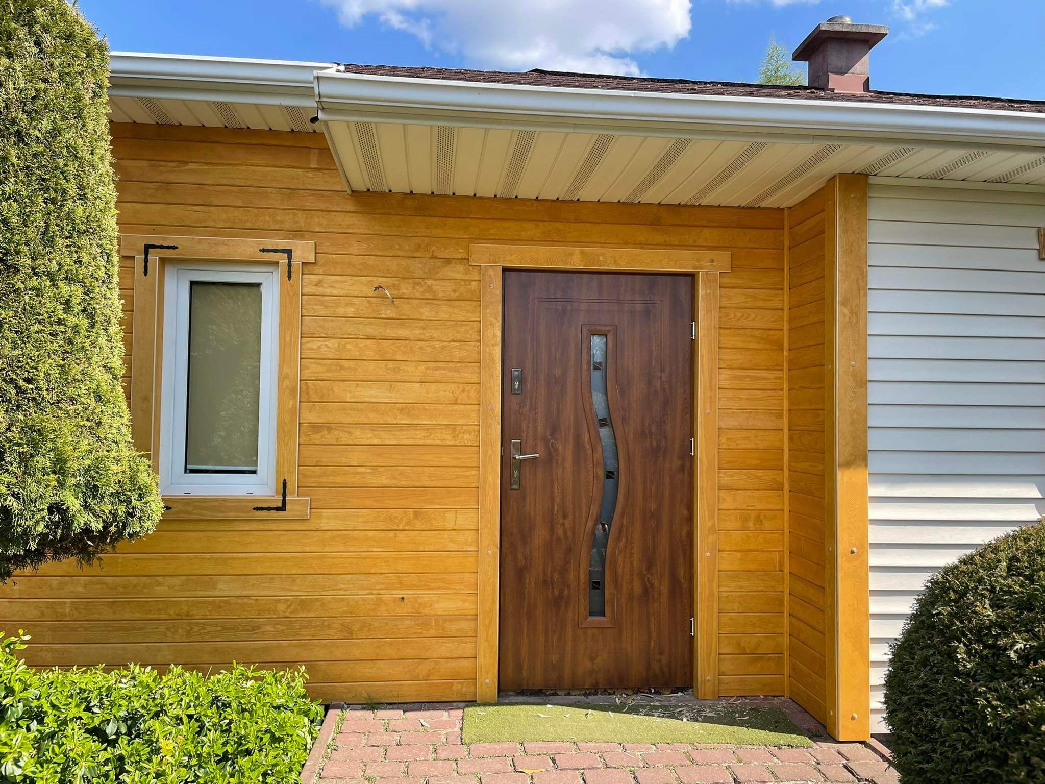 Żółta drewniana elewacja budynku z brązowymi drzwiami wejściowymi z przeszkleniem o falistym kształcie, małe okno z ozdobnymi narożnikami i zielony trawnik przed wejściem.