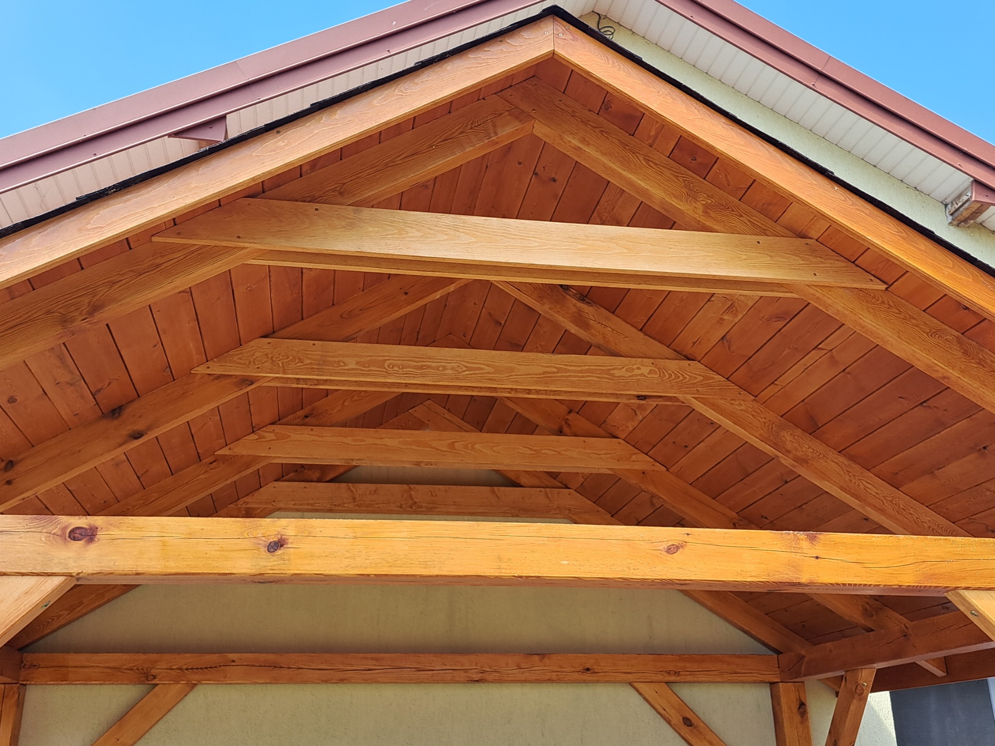 Drewniana konstrukcja dachu altany z widocznymi belkami, deskami i krokwiami, wykończona brązową blachą dachową i białymi panelami, na tle błękitnego nieba.