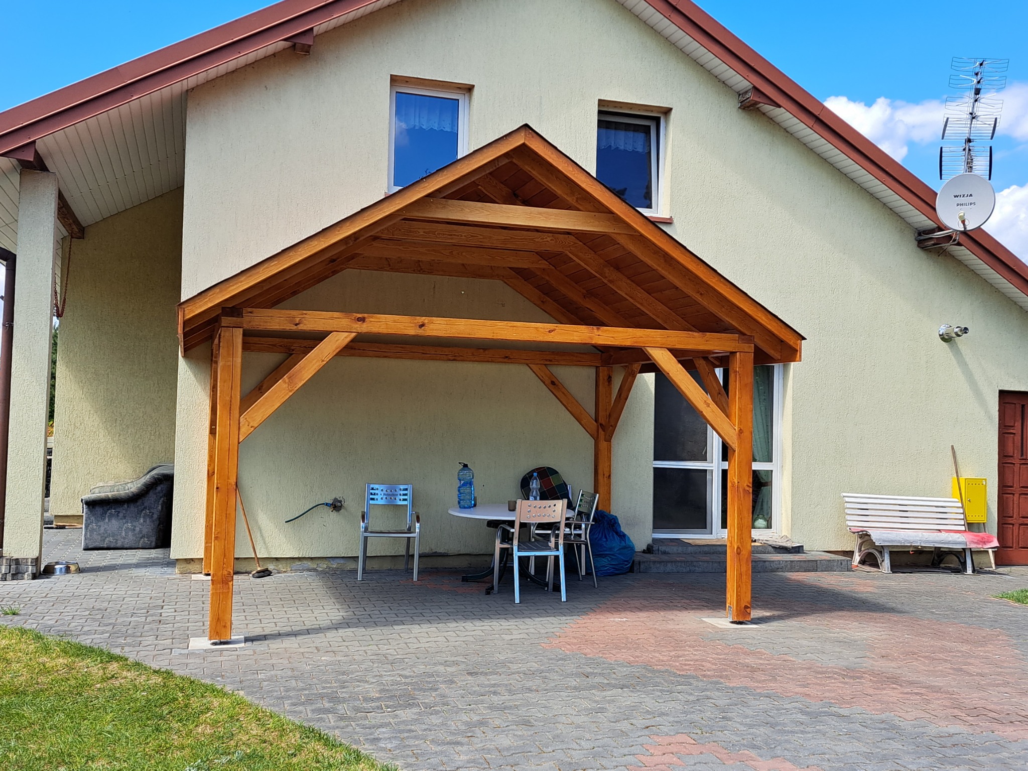 Drewniana konstrukcja zadaszenia tarasu przy domu jednorodzinnym, widok na stół z krzesłami i butelkami wody, kostka brukowa na podłożu, antena satelitarna na ścianie domu.