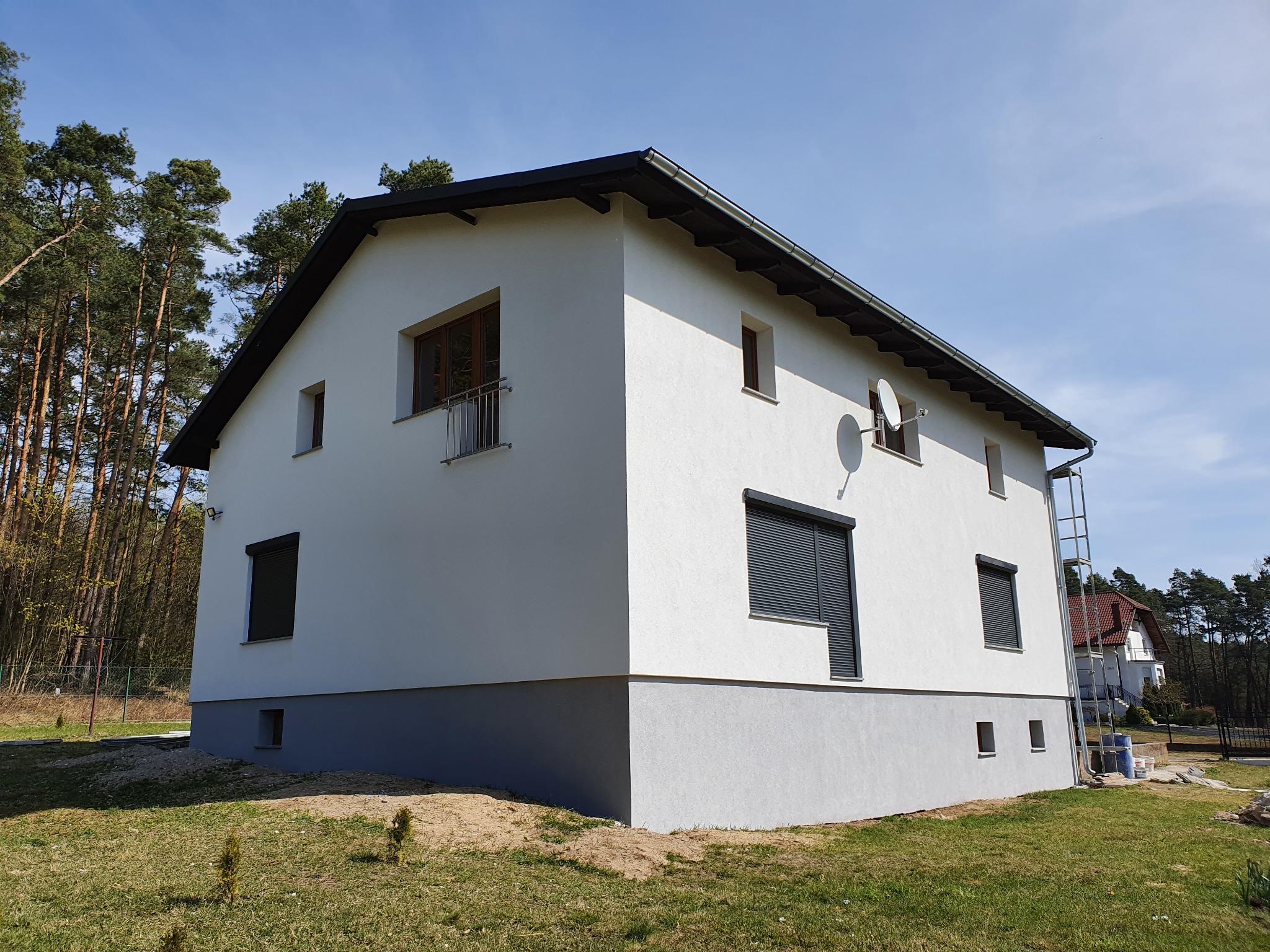 Dwukondygnacyjny dom z białą elewacją i szarym cokołem, widoczny z perspektywy ogrodu, z fragmentem lasu w tle i anteną satelitarną na ścianie.