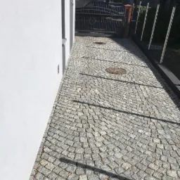 Ułożona szara nawierzchnia z kostki, widoczne studzienki kanalizacyjne i brama wjazdowa, obok biała ściana budynku.