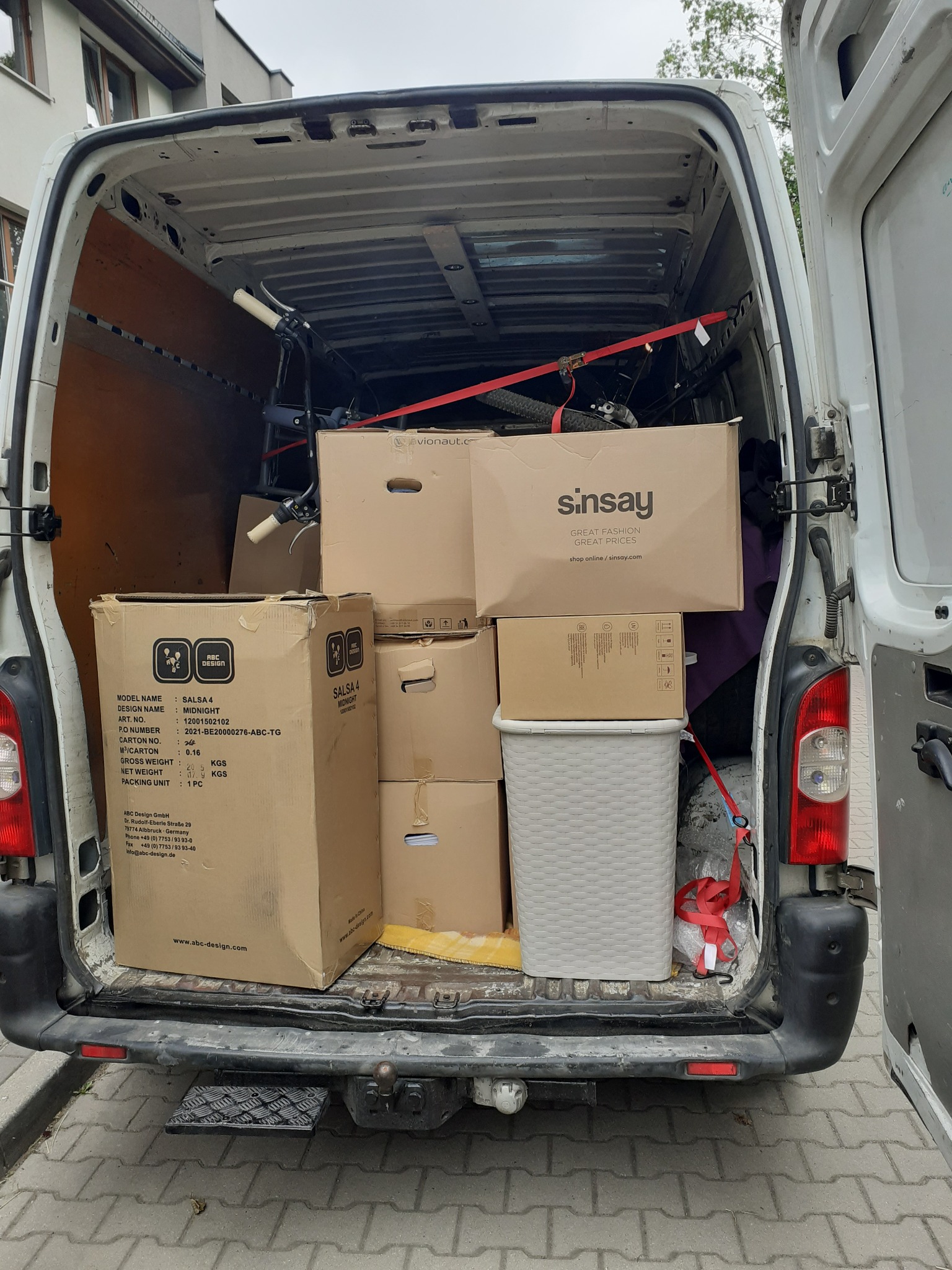 Wnętrze białego busa wypełnione kartonami różnych rozmiarów, w tym karton z logo Sinsay i ABC Design, zabezpieczone czerwonymi pasami, widoczny rower i kosz na pranie.